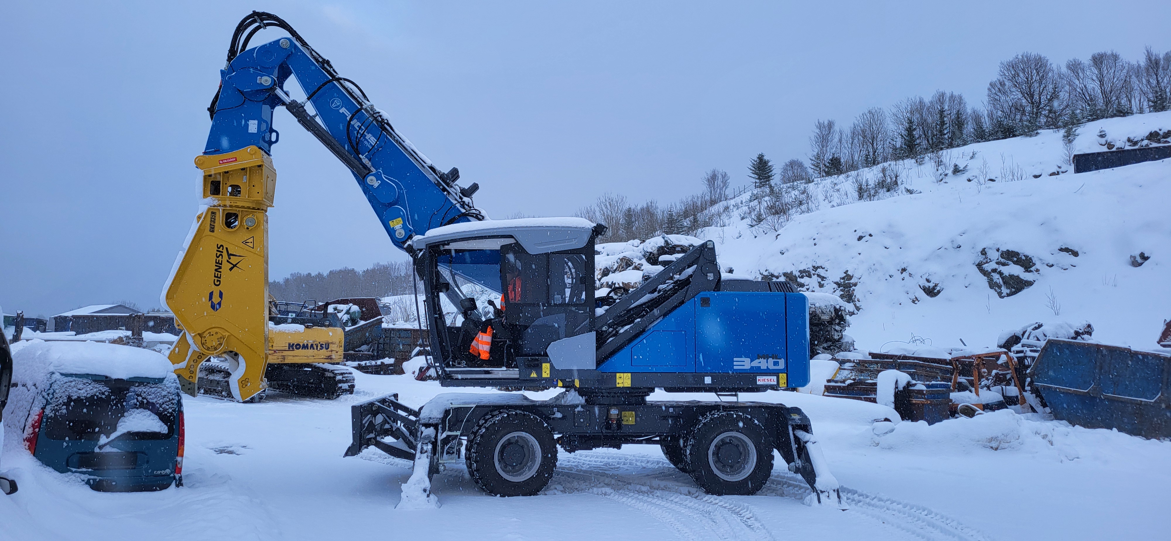Blå Fuchs MHL 340 FQC med gul Genesis saks, foran hvit himmel og over hvit bakk full av snø 