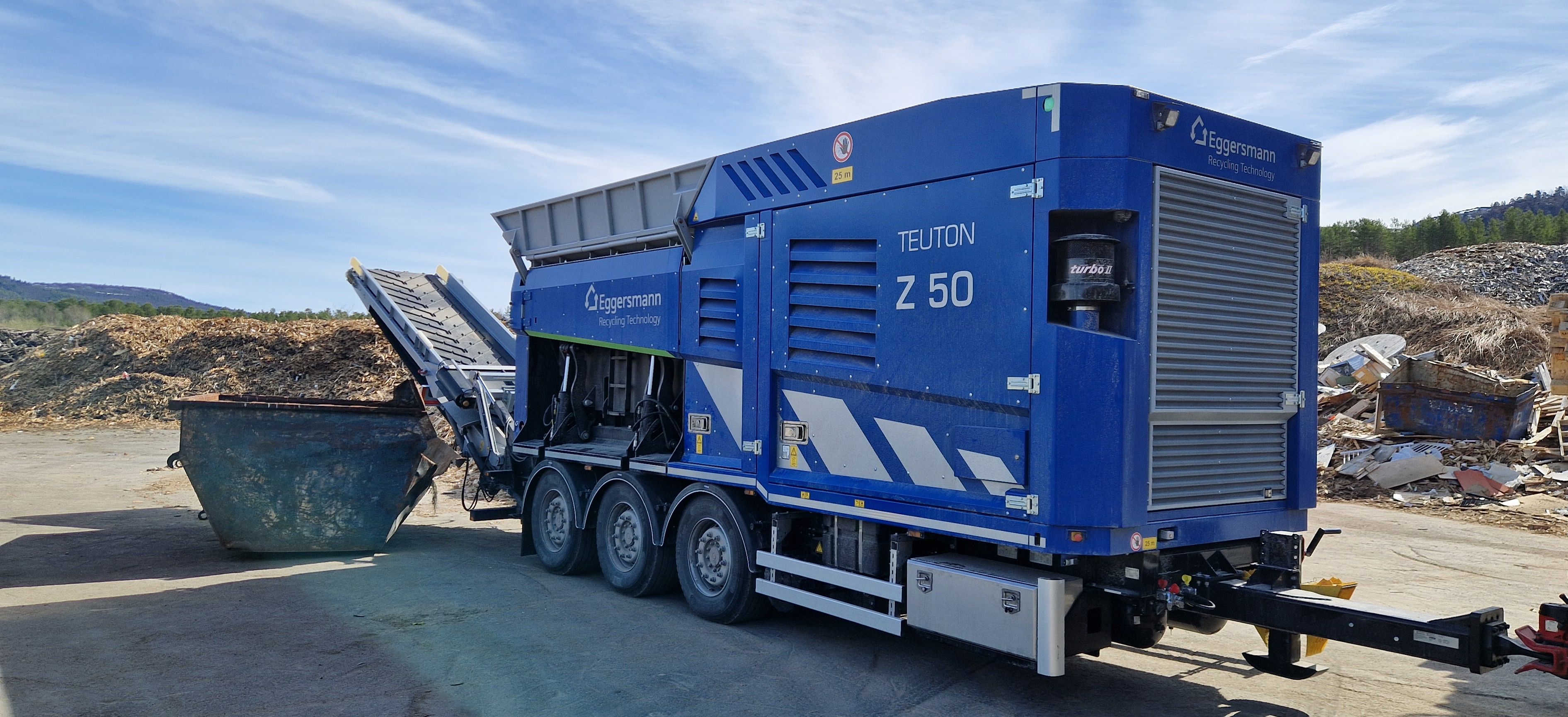 Blå Teuton kvern i bruk med container ved enden. Blå himmel og beige underlag, utendørs